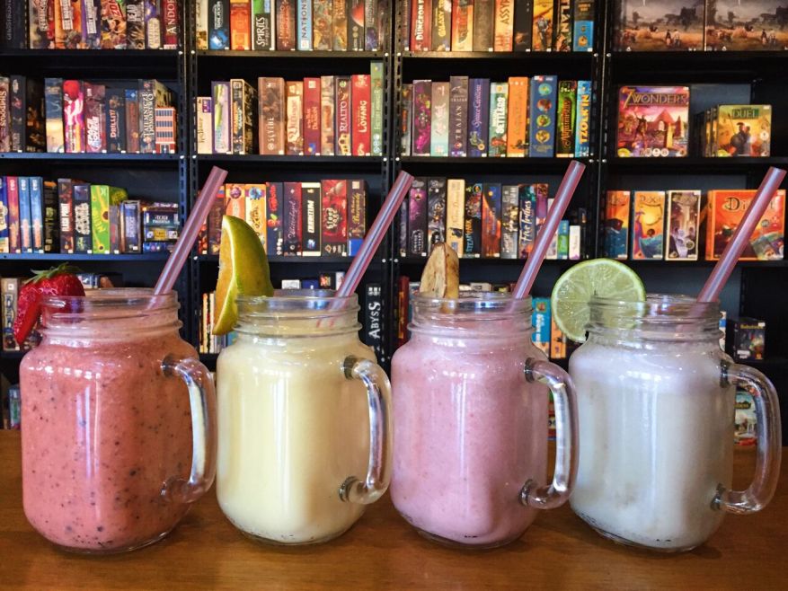 Nossos Smoothies refrescantes com iogurte, mel e frutas frescas, e, ao fundo, o acervo com cerca de 300 jogos de tabuleiro modernos